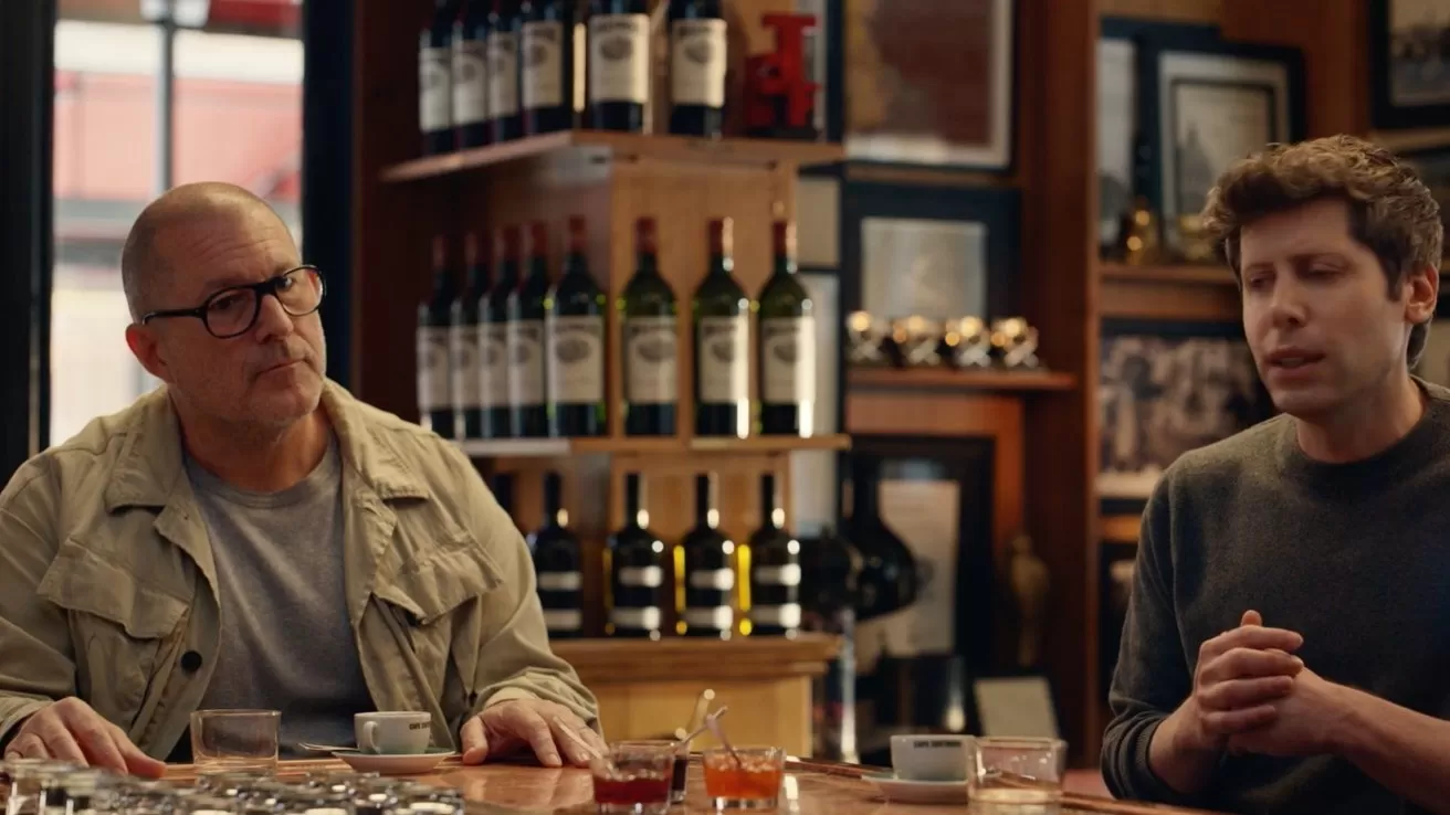 Two men sit at a bar counter talking, with wine bottles and framed photos on shelves behind them, small glasses and dishes spread out on the wooden countertop in front.