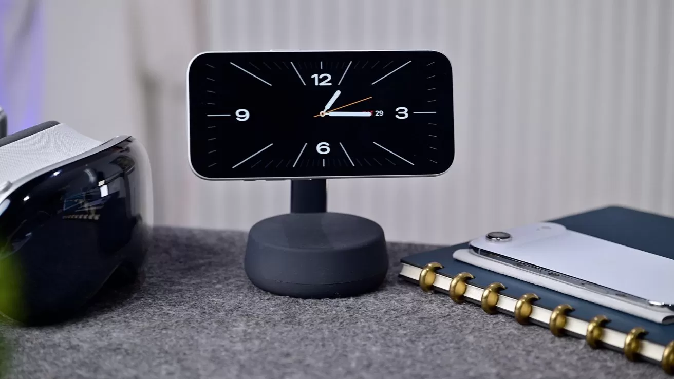 Small digital clock on a stand showing analog-style time on a desk, beside a VR headset, notebook, and pen, against a softly lit background