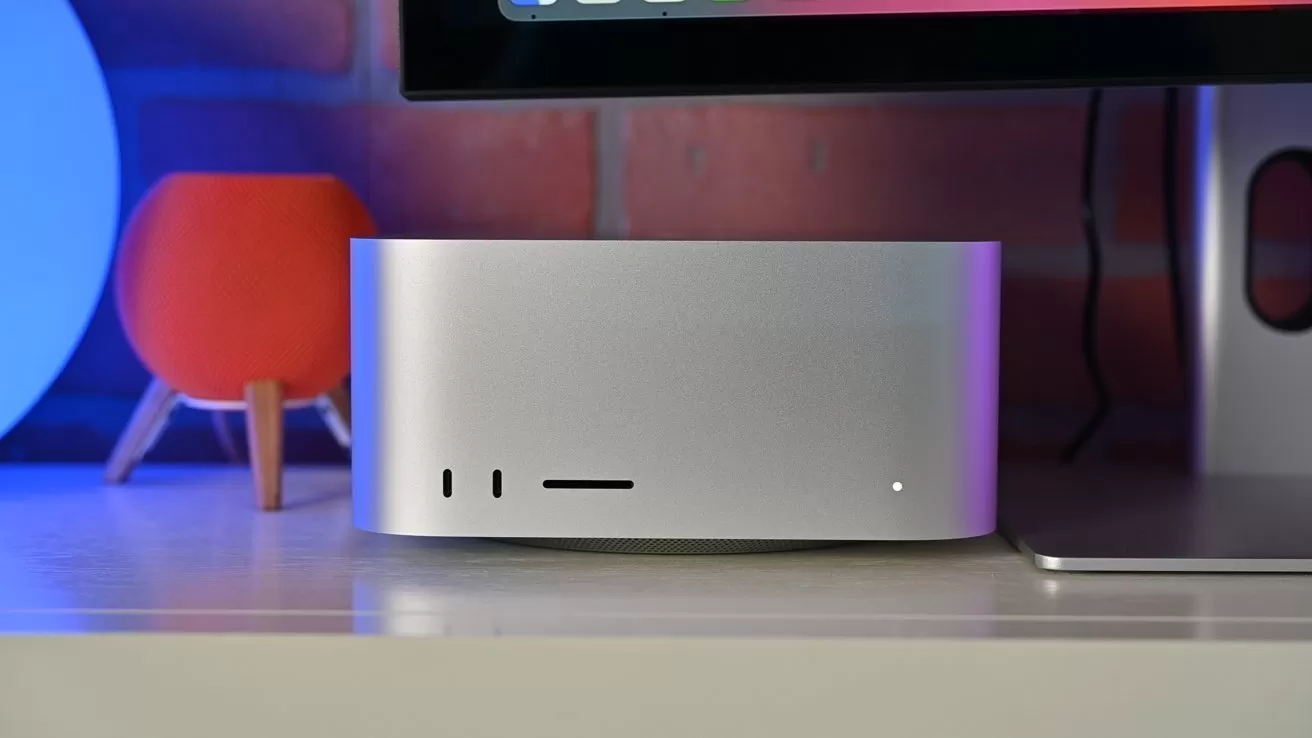Silver desktop computer with front ports and tiny status light, sitting beneath a monitor on a desk, with a small red smart speaker and brick wall background softly lit in blue and purple