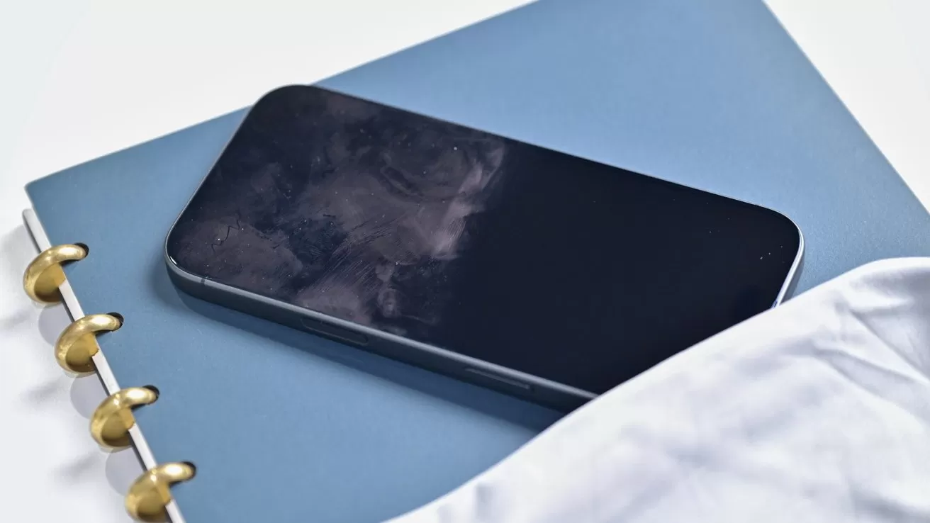 Smartphone with fingerprint smudges on a dark screen resting on a closed blue notebook with gold rings, partially covered by a white cloth or bedsheet