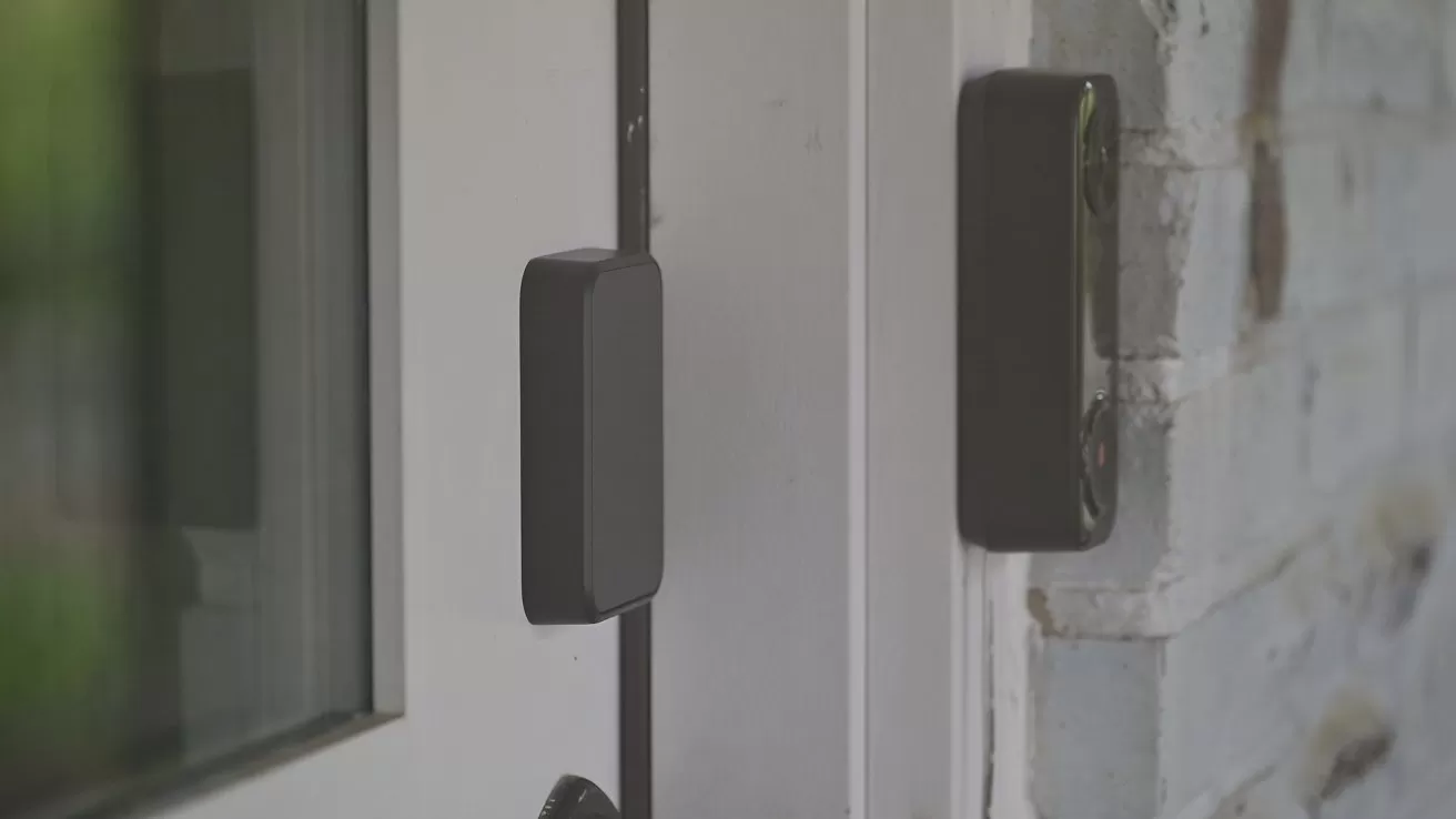 Close-up of a closed glass door with a rectangular smart sensor or contact sensor mounted on the door frame and a matching piece on the adjacent brick wall