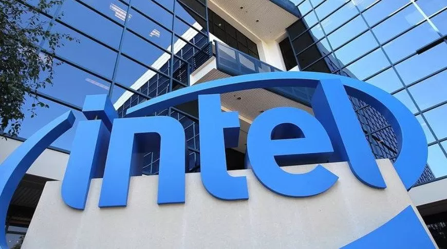 Large blue Intel logo in front of a modern glass office building, with reflective windows and metal framing under a clear sky