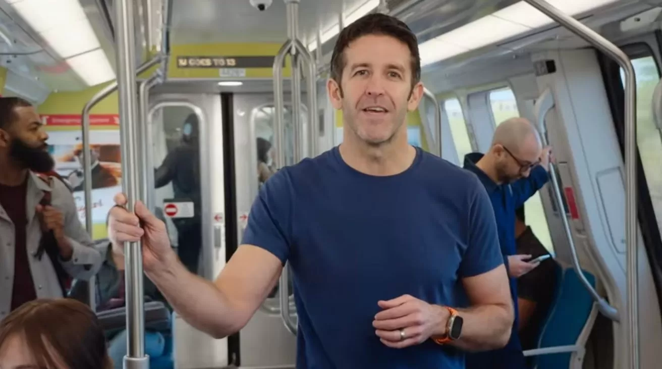 Man in blue T-shirt stands holding a subway pole, looking toward the camera, while other passengers ride behind him, some seated, some standing, one using a smartphone.