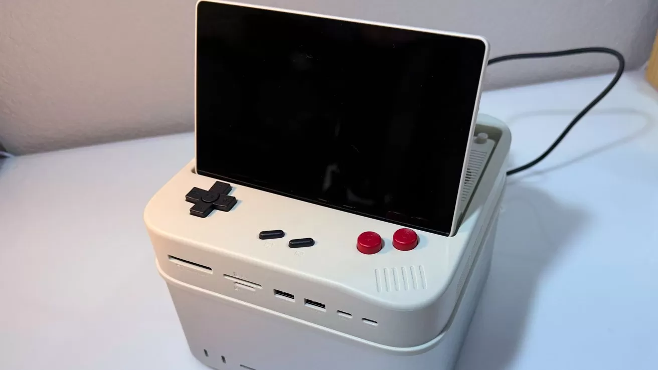 Small retro-style gaming console with a large black screen, white base, black D-pad, two red buttons, and connected power cable, sitting on a white surface near a wall.