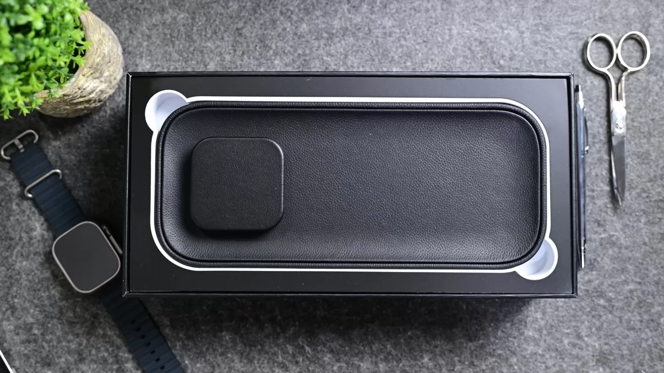 Black smartwatch packaging box on a gray surface, with watchband and watch nearby, small plant at upper left, and silver scissors at upper right.
