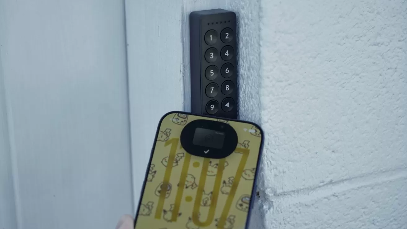 Hand holding a colorful yellow phone case near a wall-mounted electronic keypad lock on a white textured wall, suggesting contactless access or interaction with the security device