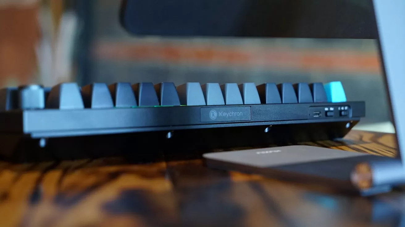 Low-angle closeup of a Keychron mechanical keyboard on a wooden desk, showing side ports and one bright blue keycap, with blurred computer equipment in the foreground and background
