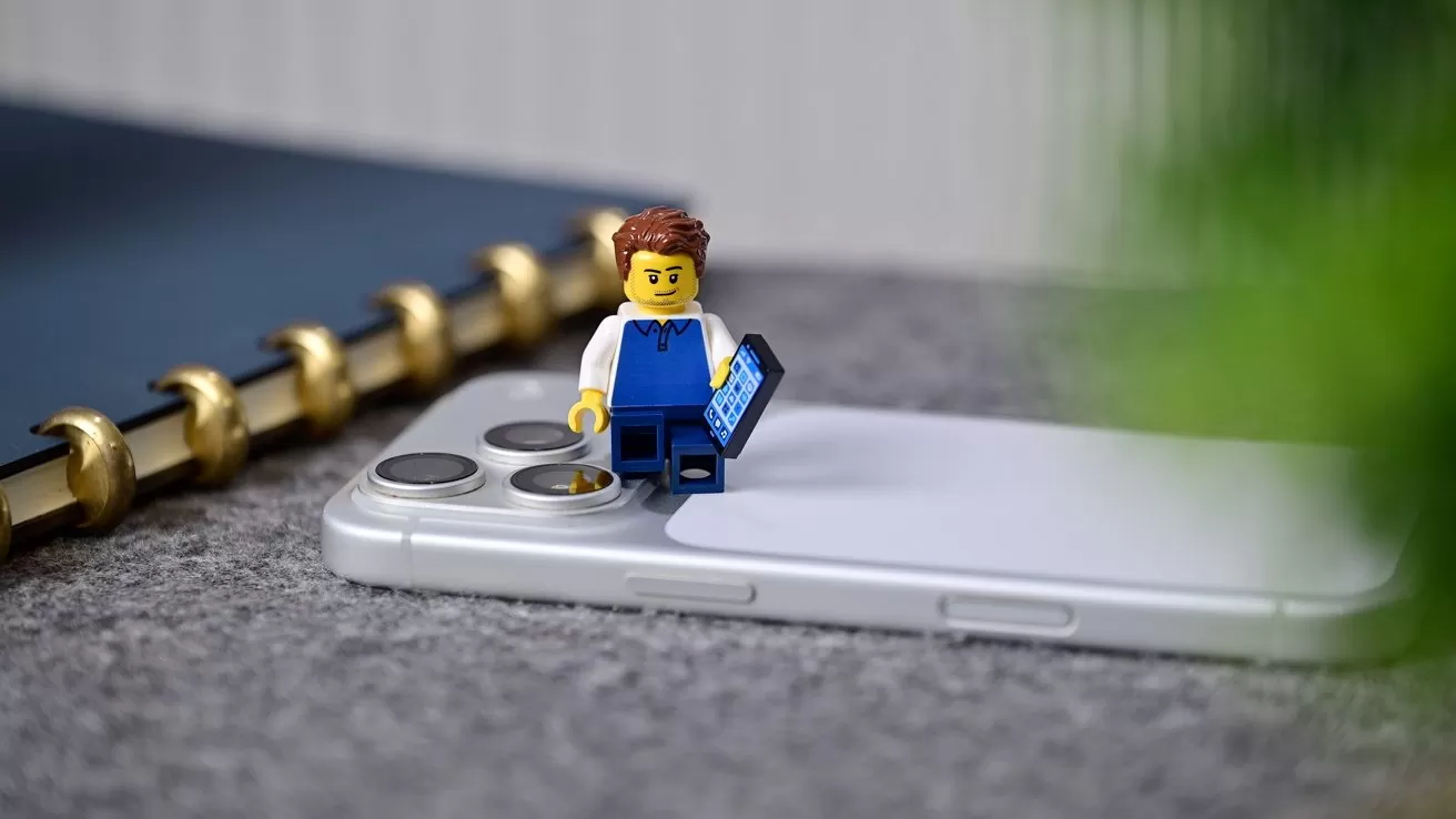 Small Lego person in blue overalls standing on a smartphone, holding a tiny laptop, with a blurred notebook and green plant in the background