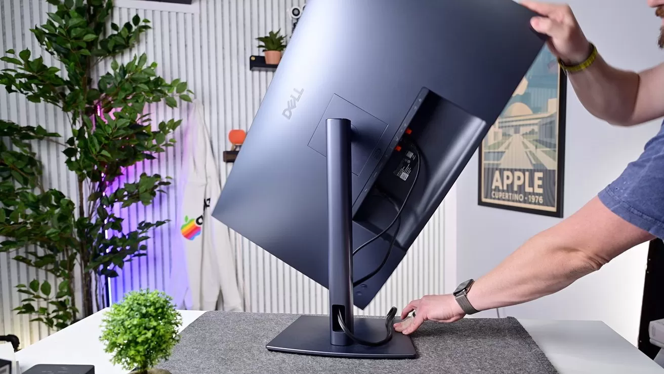 Person adjusting a large Dell monitor on a stand atop a gray desk mat, in a modern office with plants, shelves, and framed wall art in the background