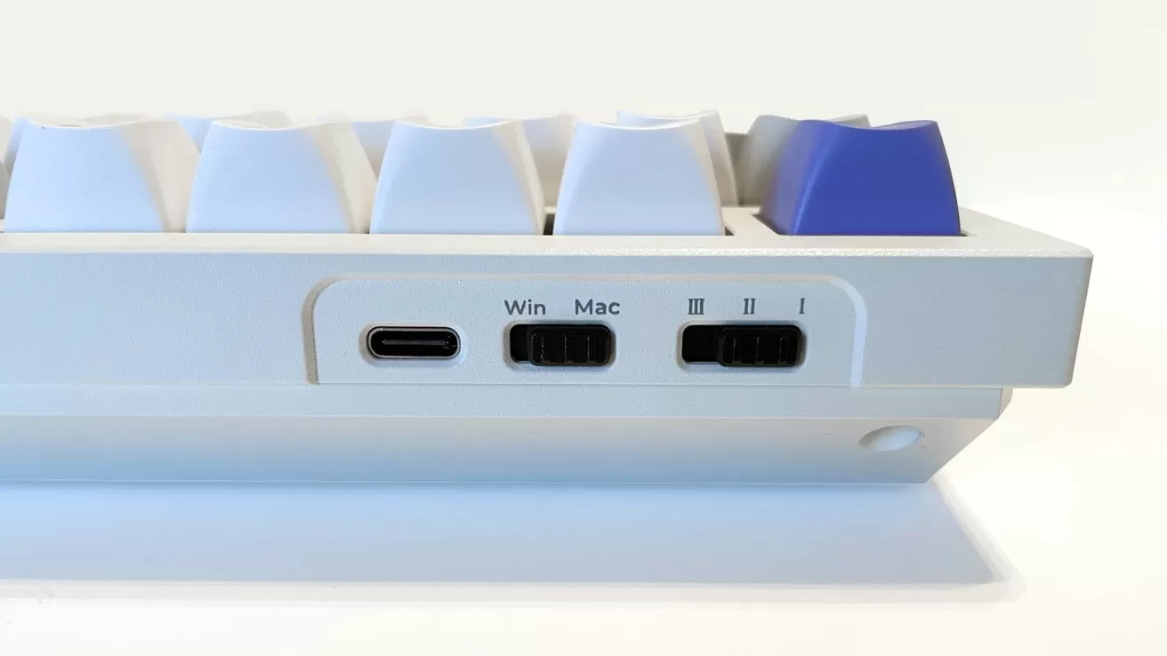 Side view of a compact mechanical keyboard showing USB-C port, Win/Mac switch, three-position toggle switch, and white keycaps with one highlighted purple key in the top right corner