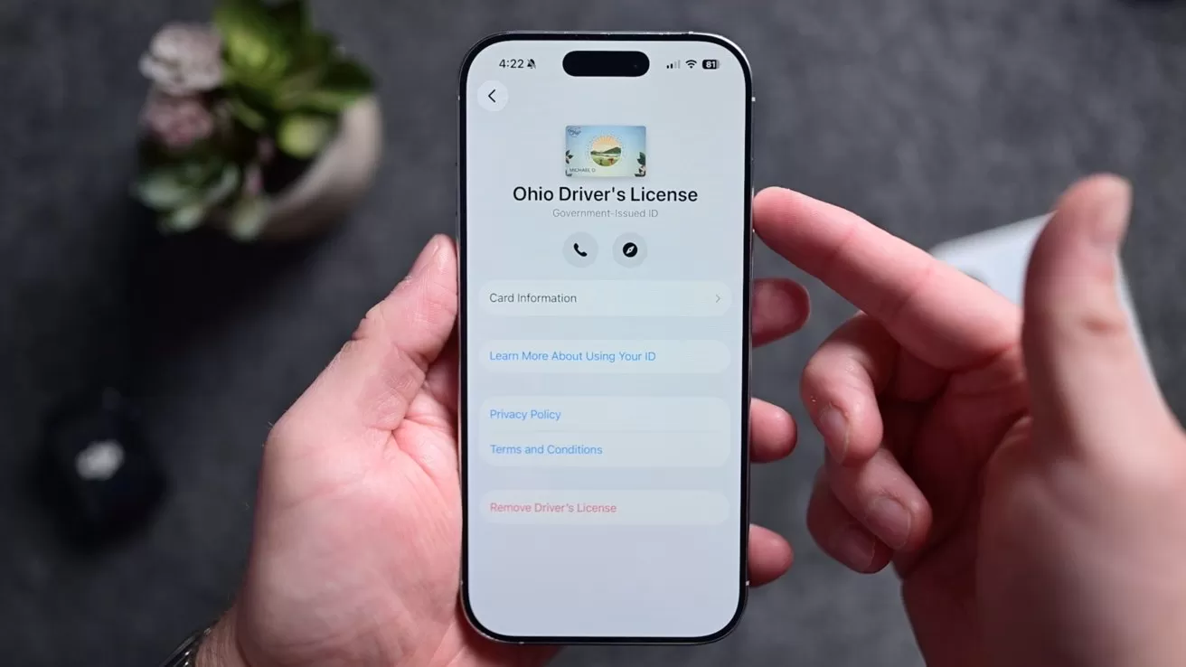 Hands holding a smartphone displaying an Ohio Driver's License app interface with user options and menu buttons; background shows blurred greenery.
