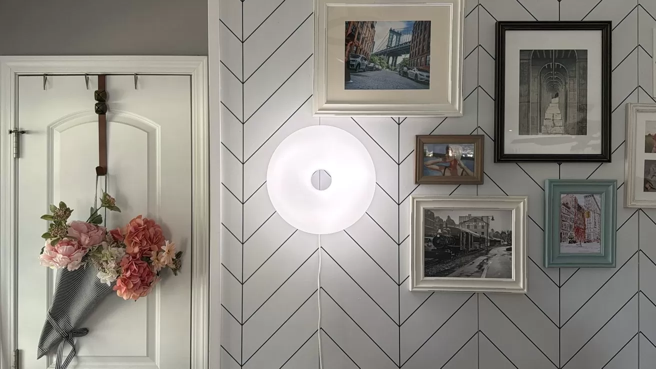 Decorative entryway with chevron-patterned wall, round glowing wall light, multiple framed city photos, and a white door on the left holding a gray umbrella filled with pink and peach flowers