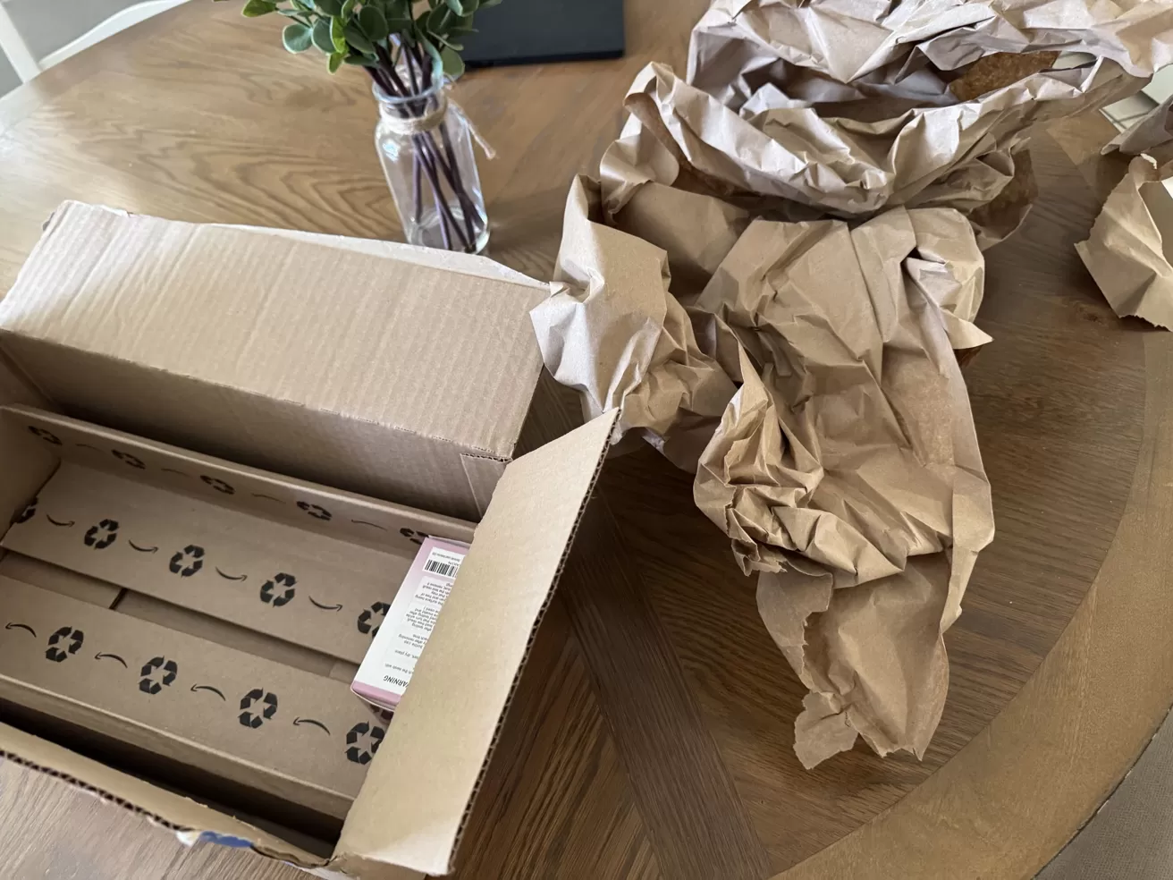 Open cardboard box on a wooden table with long packaged items inside, crumpled brown packing paper beside it, and a small glass vase holding green leafy stems in the background