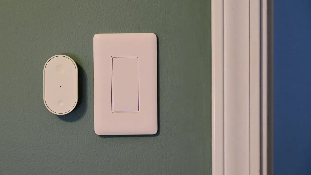 Green wall with a modern rectangular white light switch and a smaller oval white smart sensor beside it, next to a white door frame on the right
