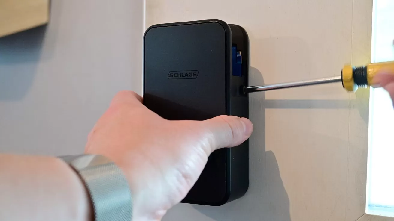 Person using a small screwdriver to tighten or loosen a screw on a rectangular black electronic device mounted on a wall, with one hand holding the device steady