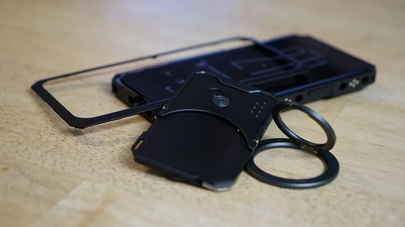 Disassembled black iPhone case with modular metal frame, backplate, and two circular attachment rings lying on a light wooden surface.