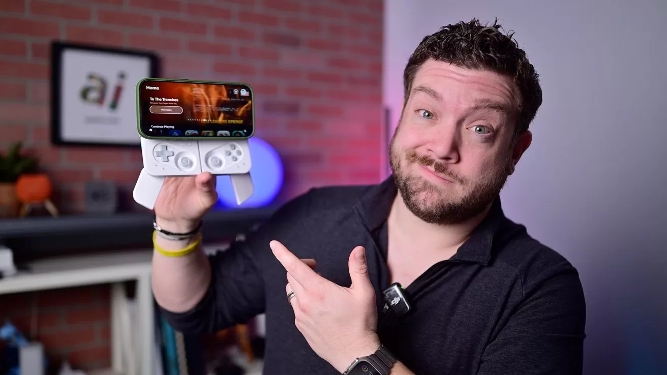 Man in dark shirt holding a smartphone with detachable game controller, smiling and pointing at it in a room with brick wall background.