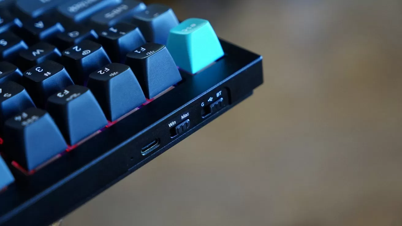 Close-up of a mechanical keyboard corner showing dark keys, one teal keycap, a USB-C port, and small switches labeled Windows, Mac, wired, and Bluetooth on the side.