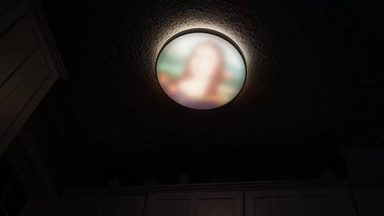 Ceiling light fixture showing a blurred circular picture with soft colors, casting a dim glow over dark cabinets and a textured ceiling in a low-lit room