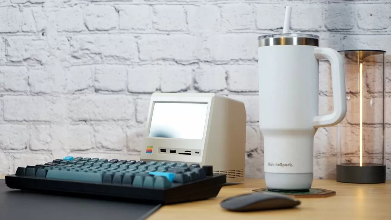 Retro-style small computer, gray keyboard, tall white tumbler with straw, glowing LED lamp on wooden table, white brick wall background.