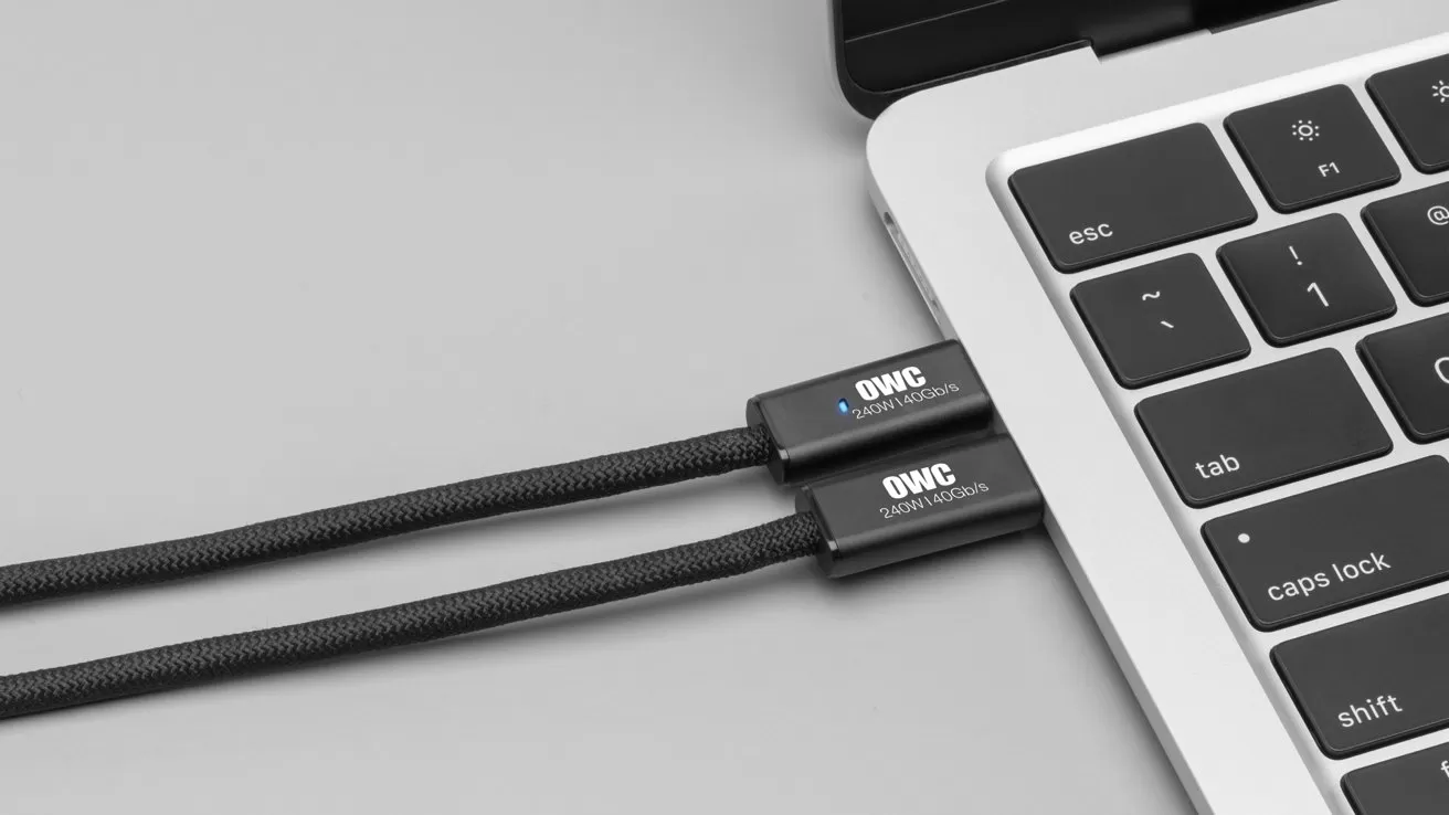 Close-up of a laptop keyboard with two black braided OWC USB-C cables plugged into adjacent ports on the side, resting on a smooth light gray surface