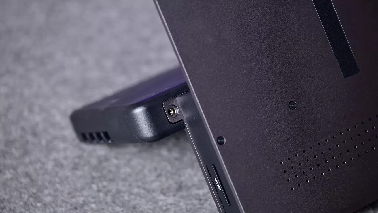 Close-up of a tablet or 2-in-1 laptop propped on its kickstand, showing side ports, ventilation holes, and a connected accessory, resting on a soft gray surface