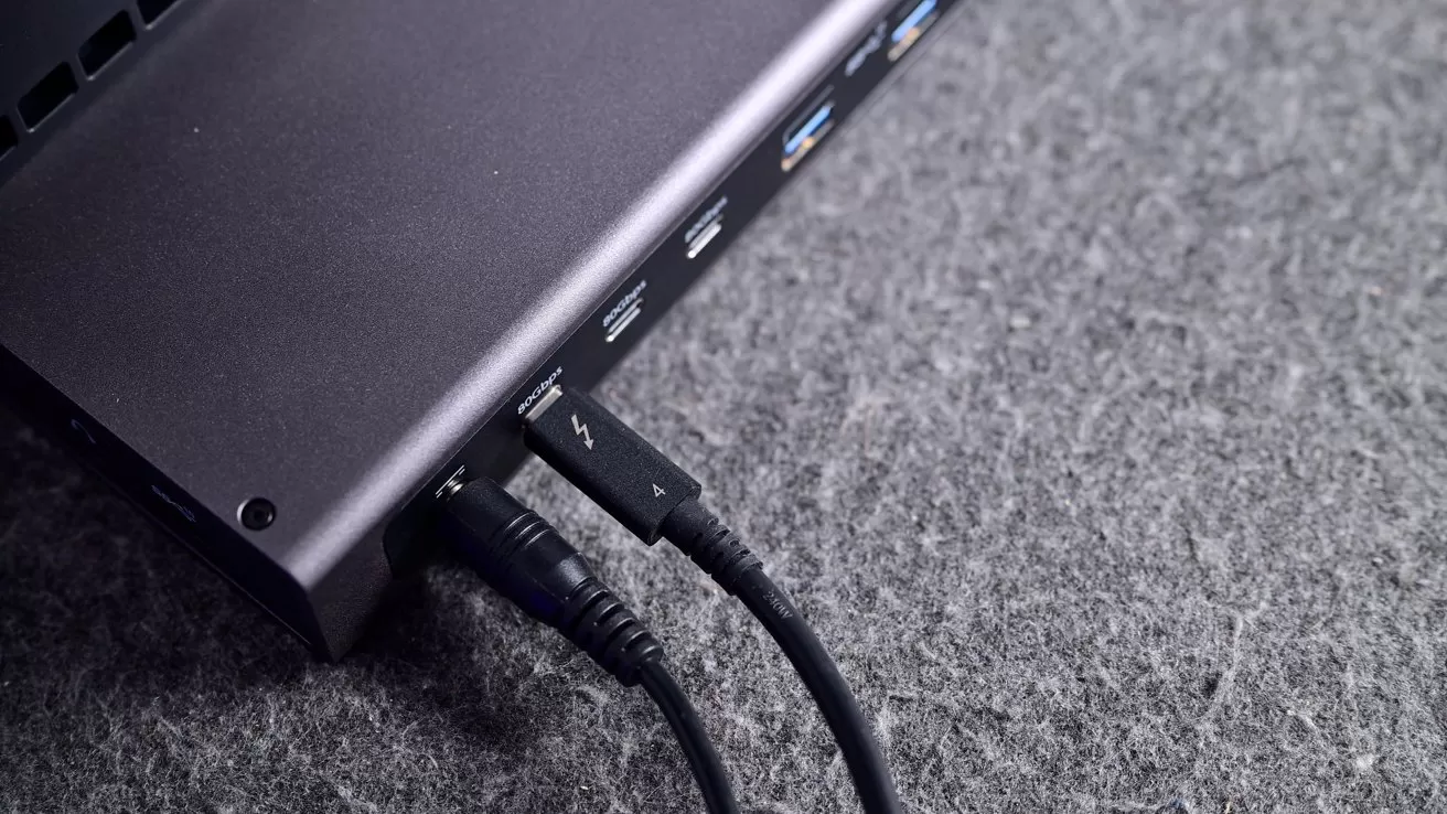 Close-up of a laptop's side ports on gray fabric, showing multiple USB ports with cables plugged in, including a Thunderbolt USB-C cable and an audio or power cable