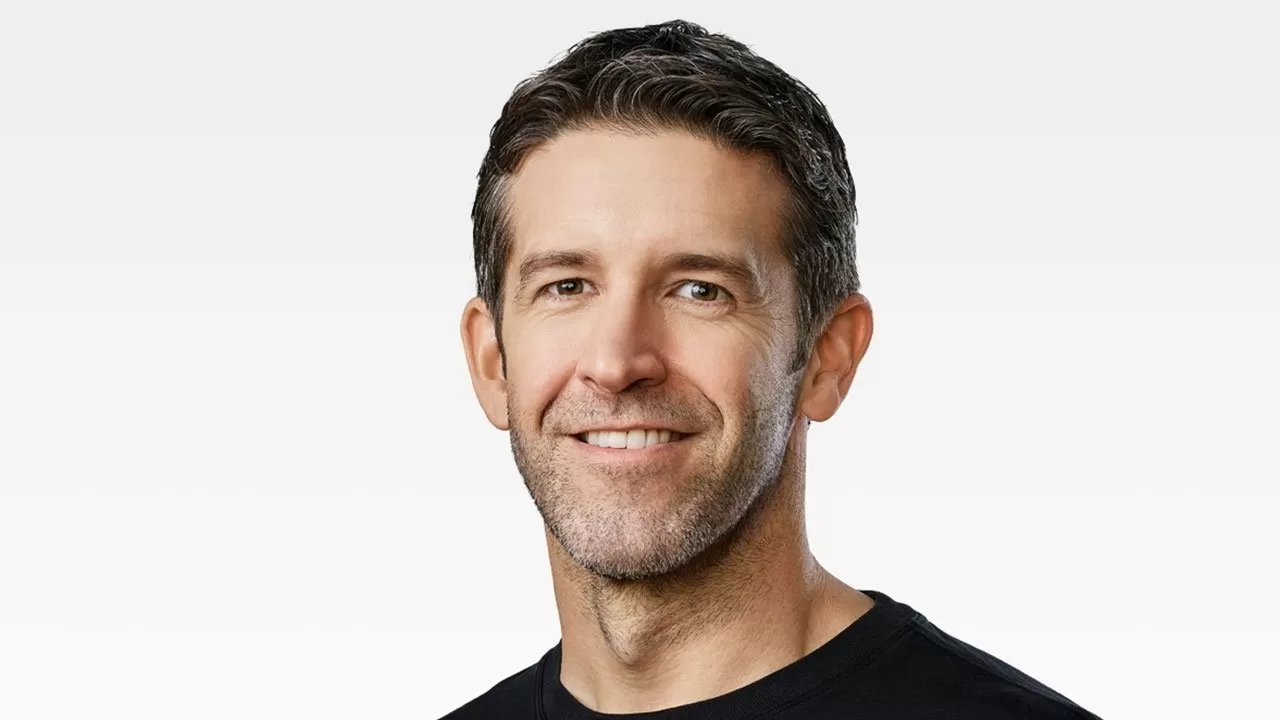 Middle-aged man with short dark hair and stubble, smiling gently, wearing a black crew-neck shirt against a plain light gray background