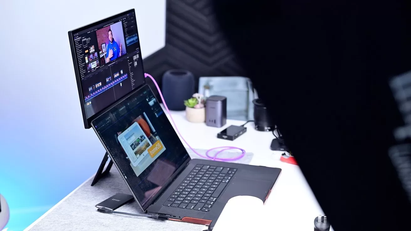 Laptop and monitor on a desk showing video editing software, surrounded by various tech accessories and a small plant.