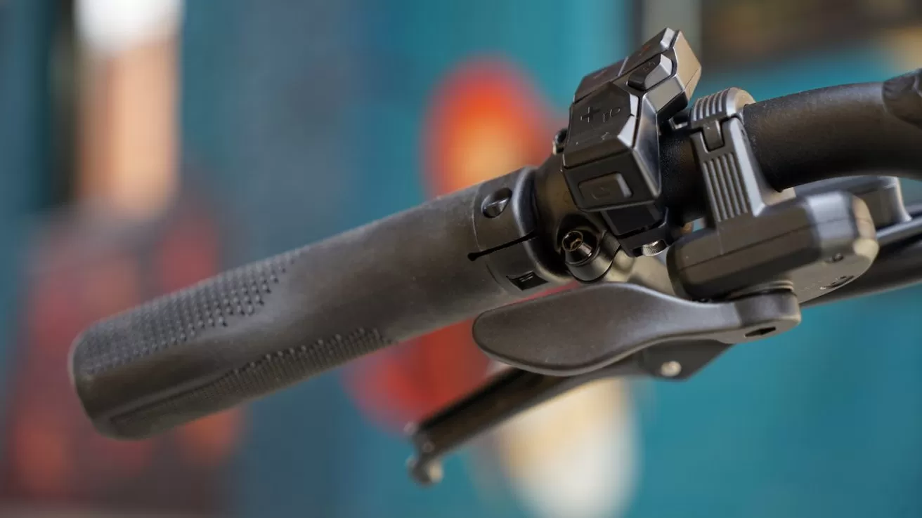 Close-up of a bicycle handlebar showing a textured rubber grip, brake lever, and electronic control buttons, with a softly blurred colorful background