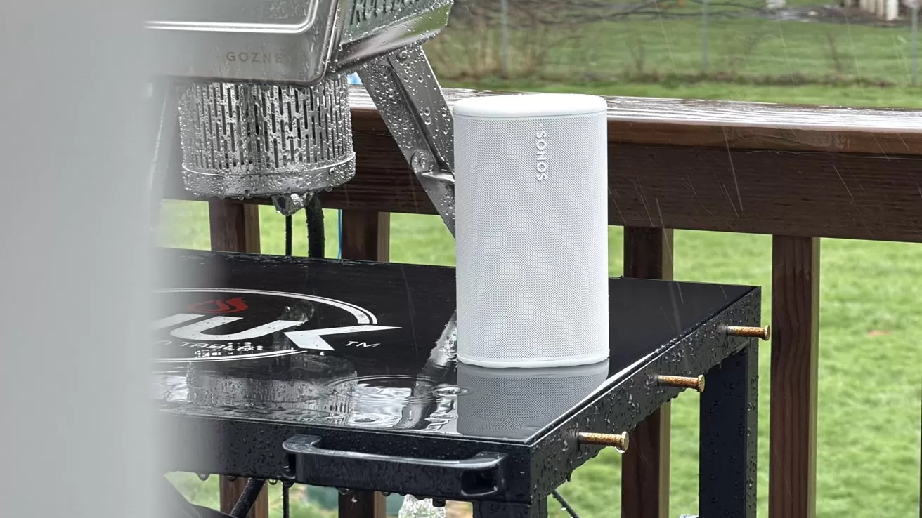 White Sonos speaker standing on a wet black outdoor grill shelf in the rain, with wooden deck railing and blurry green yard in the background