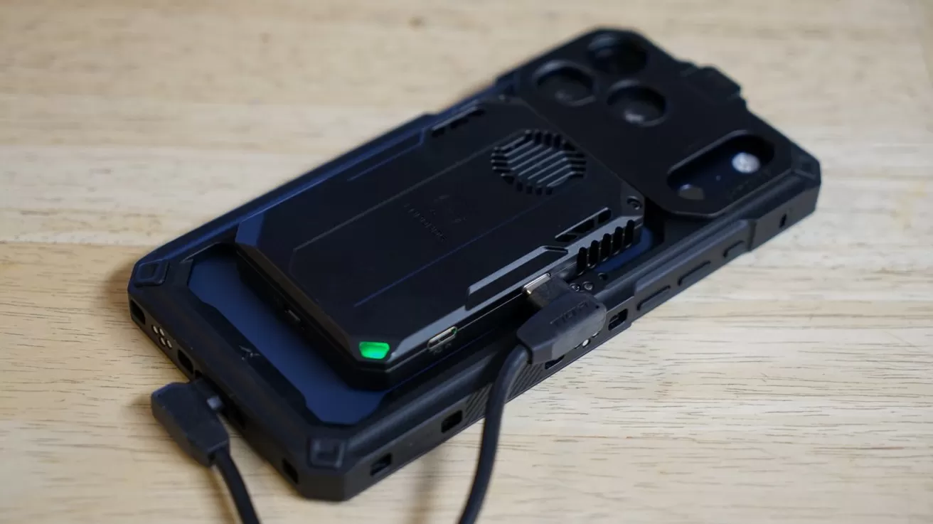 iPhone lying face down on a wooden surface, with a black external drive magnetically attached to its back and connected by a short USB cable