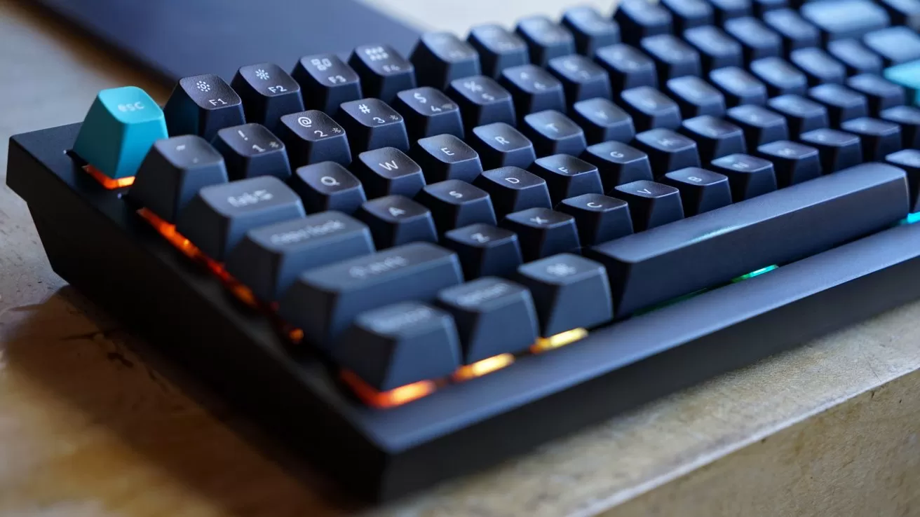 Close-up of a black mechanical keyboard with raised keycaps, teal escape key, and warm orange backlighting, sitting on a wooden desk.