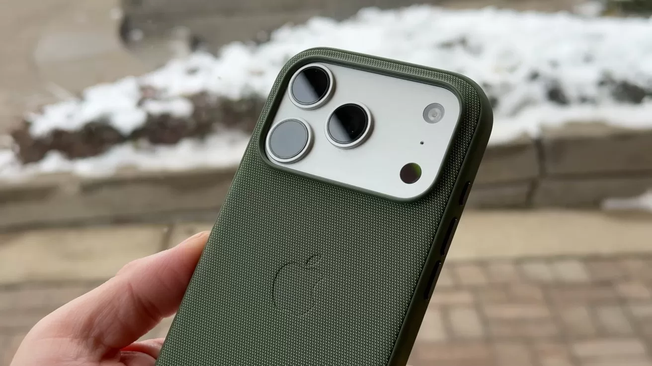 Hand holding an iPhone with a green textured Apple case, showing dual rear cameras and flash, outdoors near a snow-covered sidewalk and stone edging