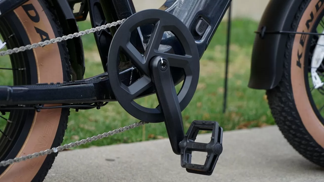 Close-up of a modern bicycle drivetrain showing black crankset, pedal, chain, and part of the frame with tan sidewall tire, parked on concrete beside green grass