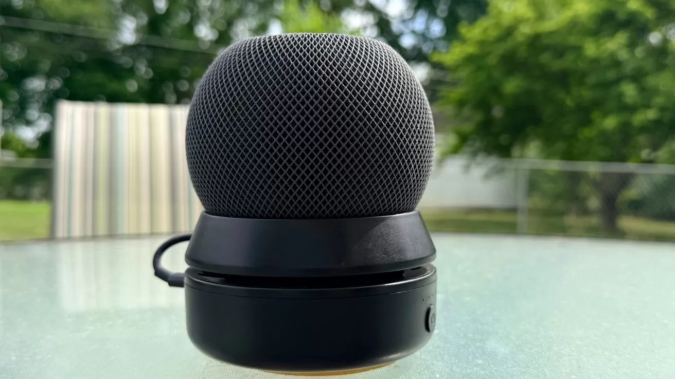 Small black spherical smart speaker on a glass outdoor table, with green trees and a striped patio umbrella softly blurred in the background