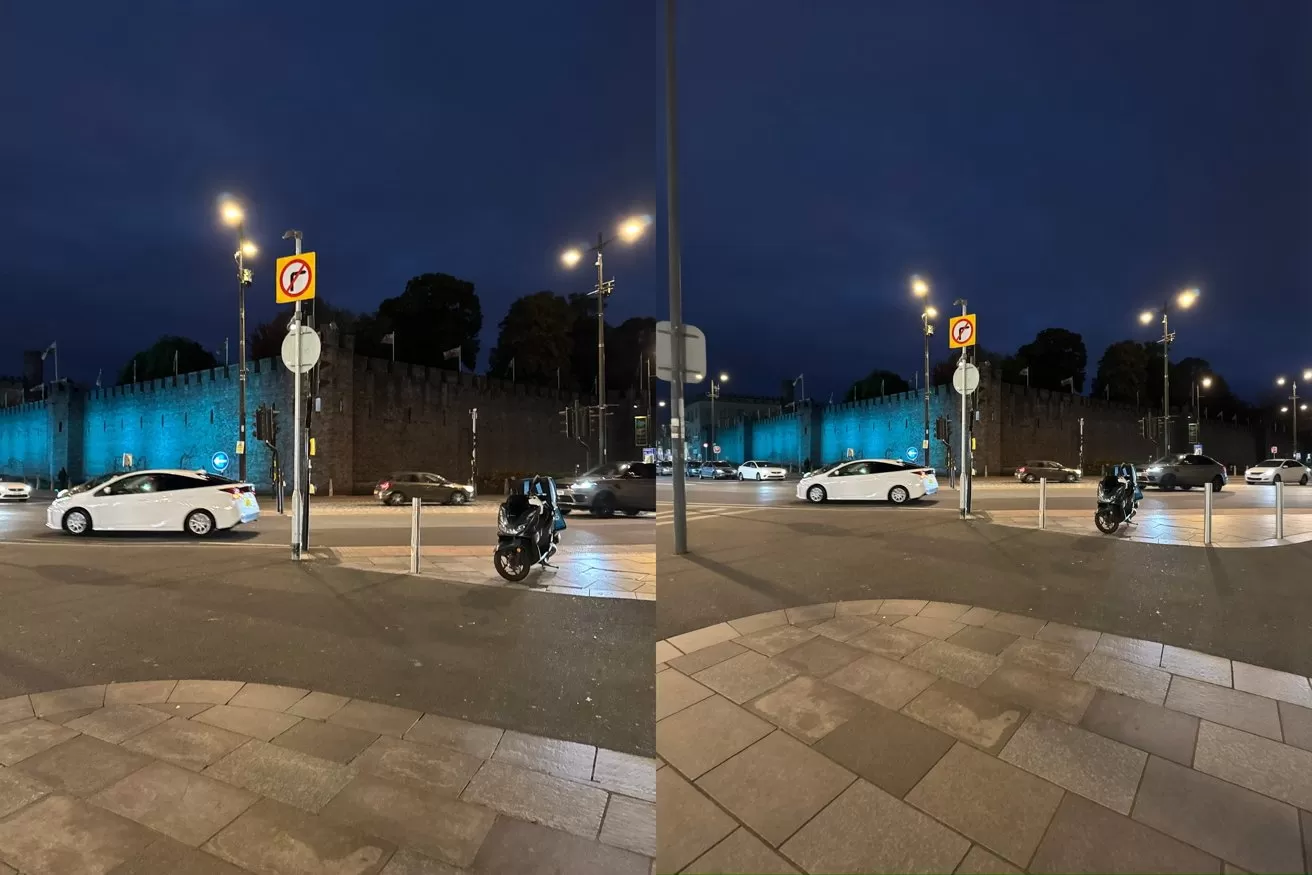 Nighttime city street with cars and motorcycles, bright streetlights, a glowing blue-lit stone wall in the background, traffic signs, and tiled sidewalk in the foreground