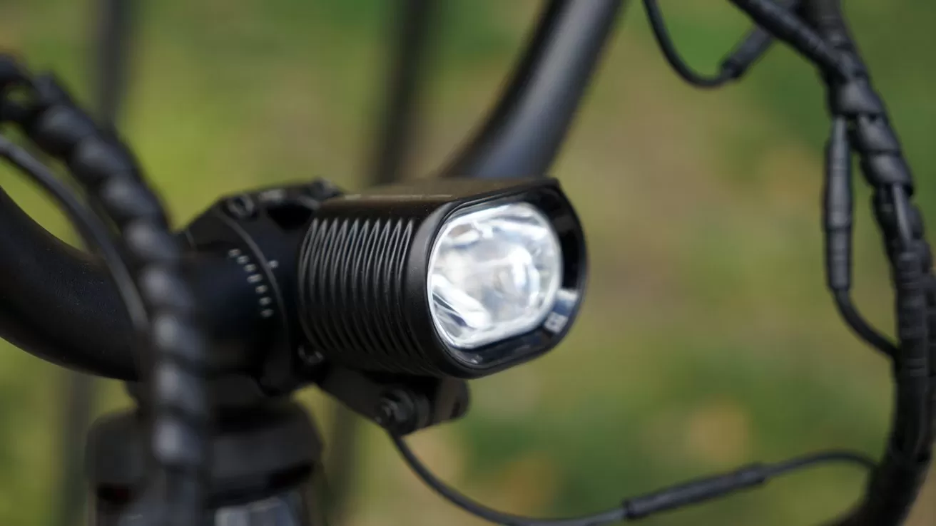 Close-up of a black bicycle handlebar-mounted headlight with ridged housing and bright lens, surrounded by coiled cables, against a blurred green outdoor background