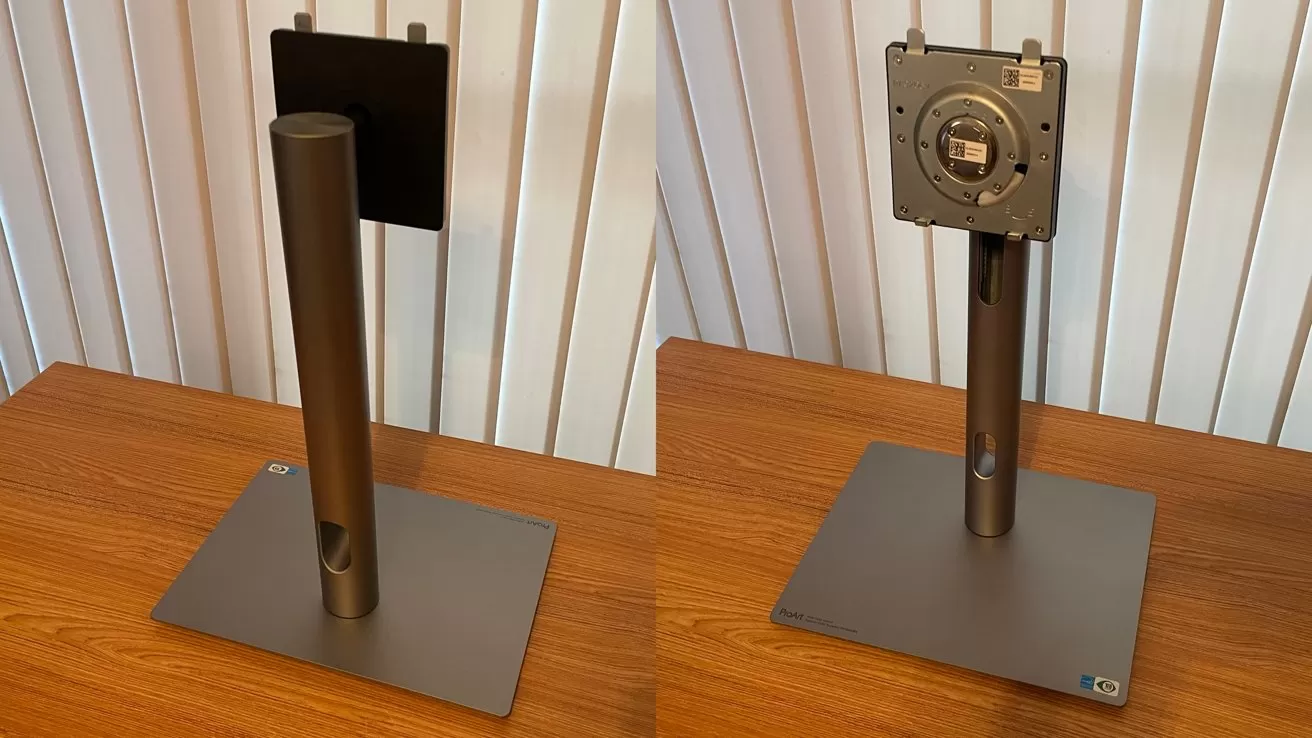 Metallic monitor stand on wooden table, featuring a square base and an adjustable vertical arm, in front of vertical blinds.