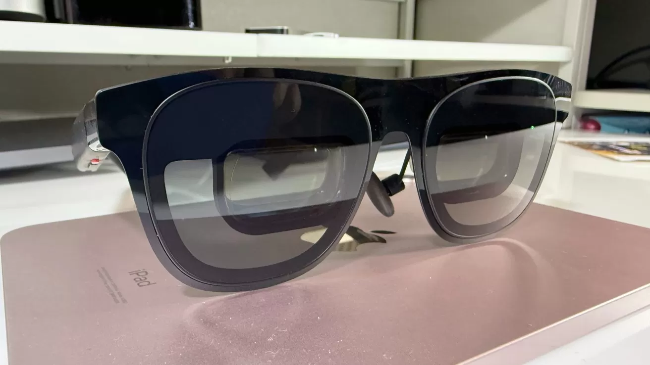 Black sunglasses with thick frames on a pink iPad, placed on a white desk.