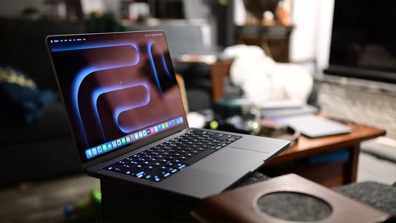 Open laptop on a living room couch armrest, glowing keyboard and abstract screensaver, with a wooden coffee table, notebooks, and blurred background furniture in soft indoor lighting
