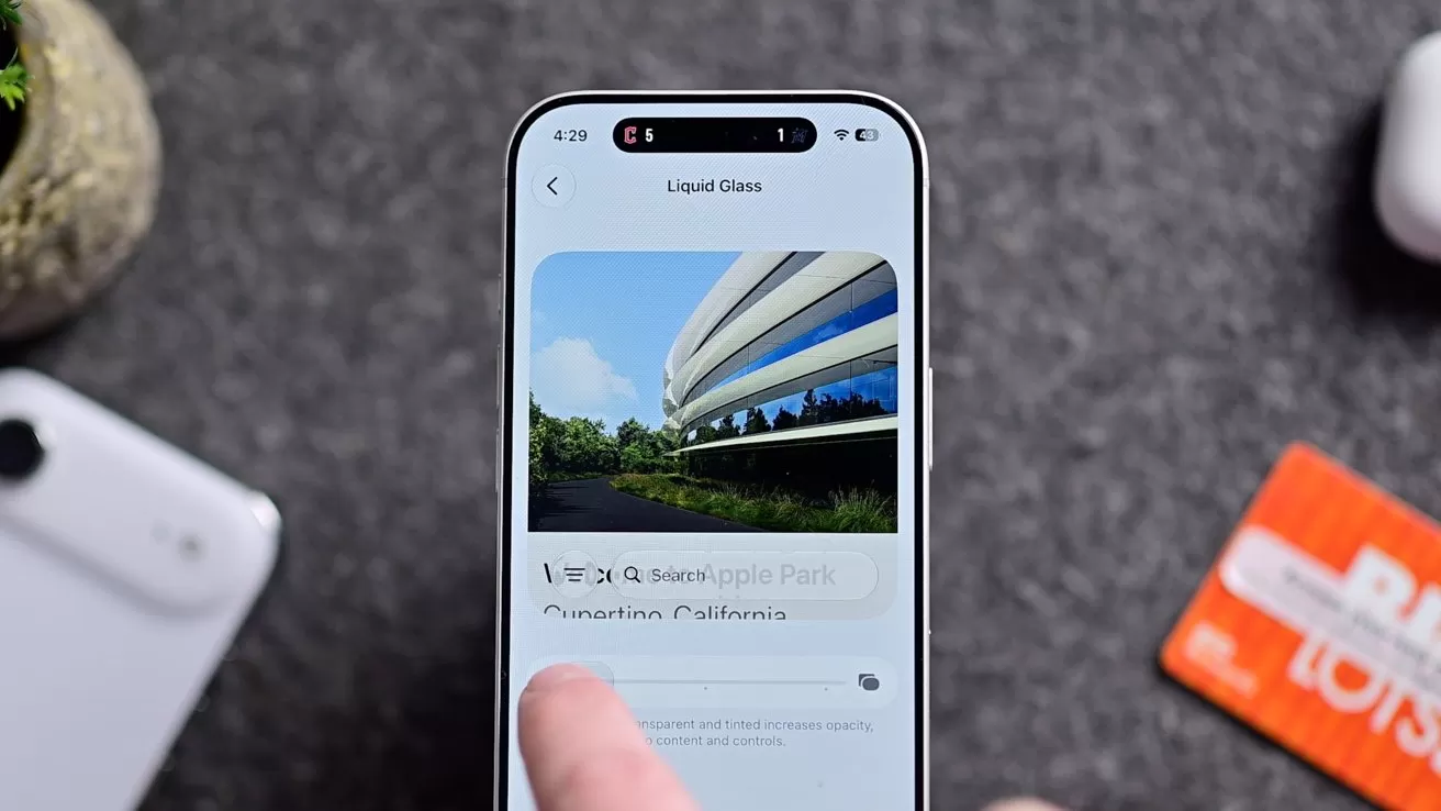Hand holding a smartphone displaying a travel or map app with a curved modern building photo, while a finger taps the screen; other gadgets and a card lie blurred in the background