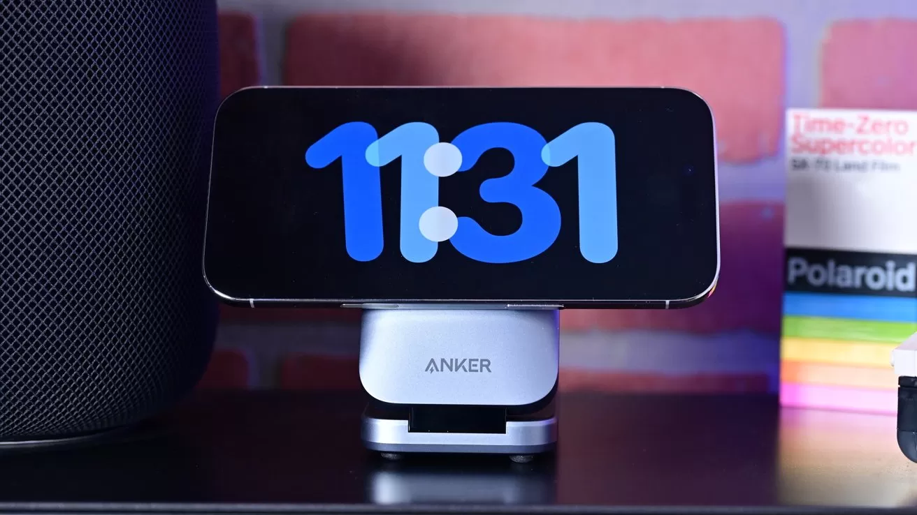 Smartphone in landscape orientation on an Anker stand displaying digital clock 11:31, placed on a desk between a large speaker and colorful Polaroid film boxes against a brick wall