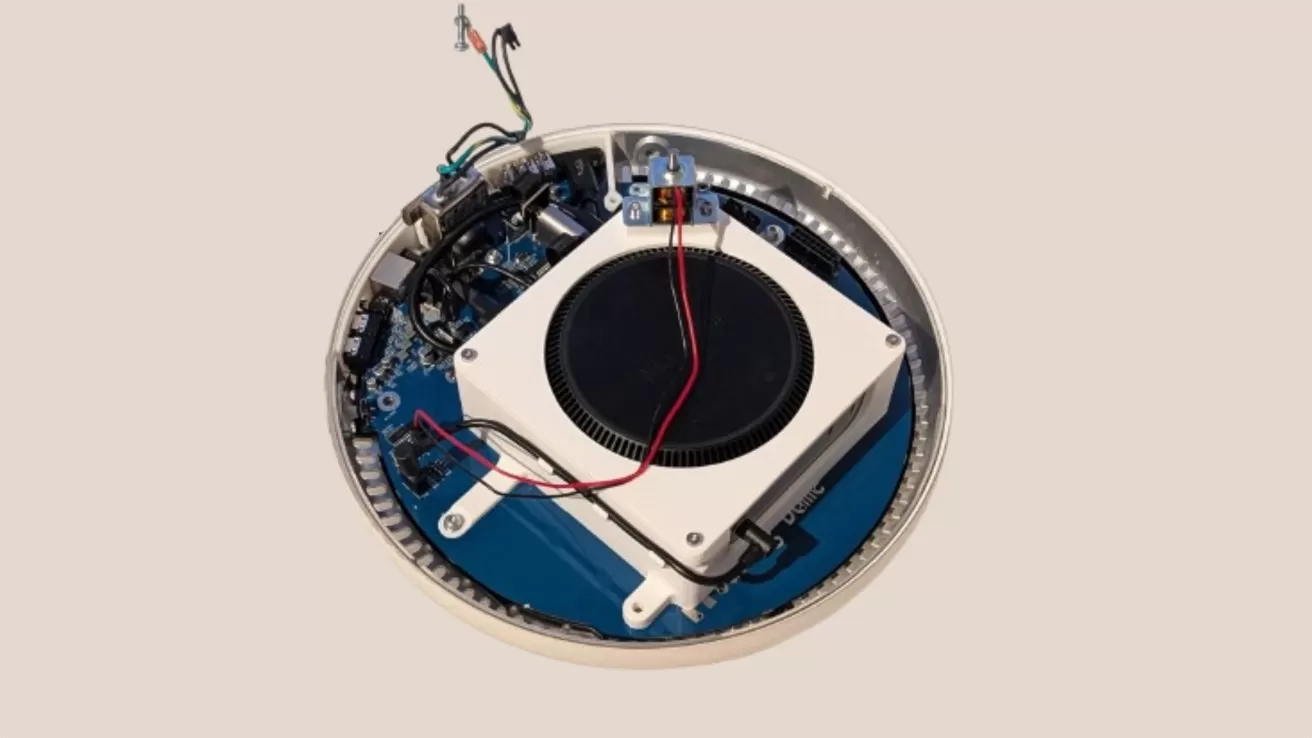 Round electronic device with its cover removed, exposing a central black circular component, circuit board, wiring, and connectors on a beige background