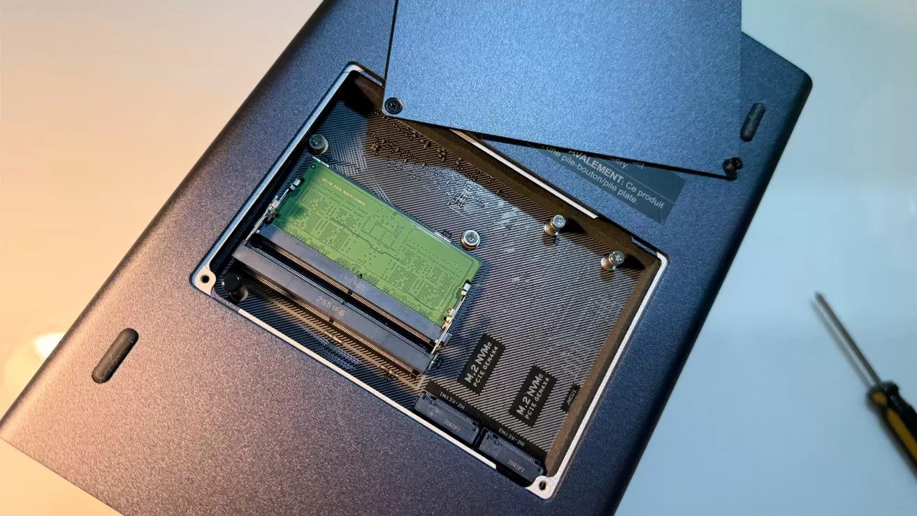 Open laptop underside showing exposed RAM slot with a single green memory module, removed access panel nearby, and a small screwdriver resting on the table beside it