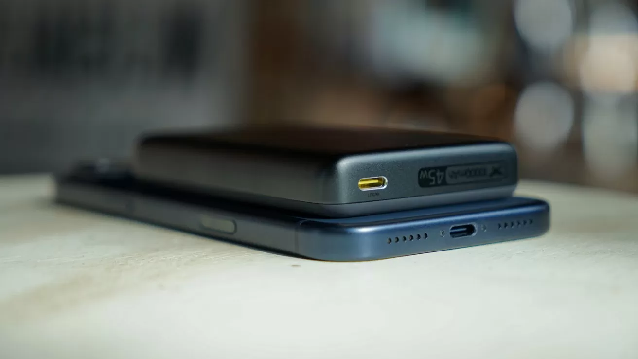 Compact black power bank resting on top of an iPhone 17 Pro Max, both lying flat on a light surface, showing their USB ports and speaker holes in soft indoor lighting