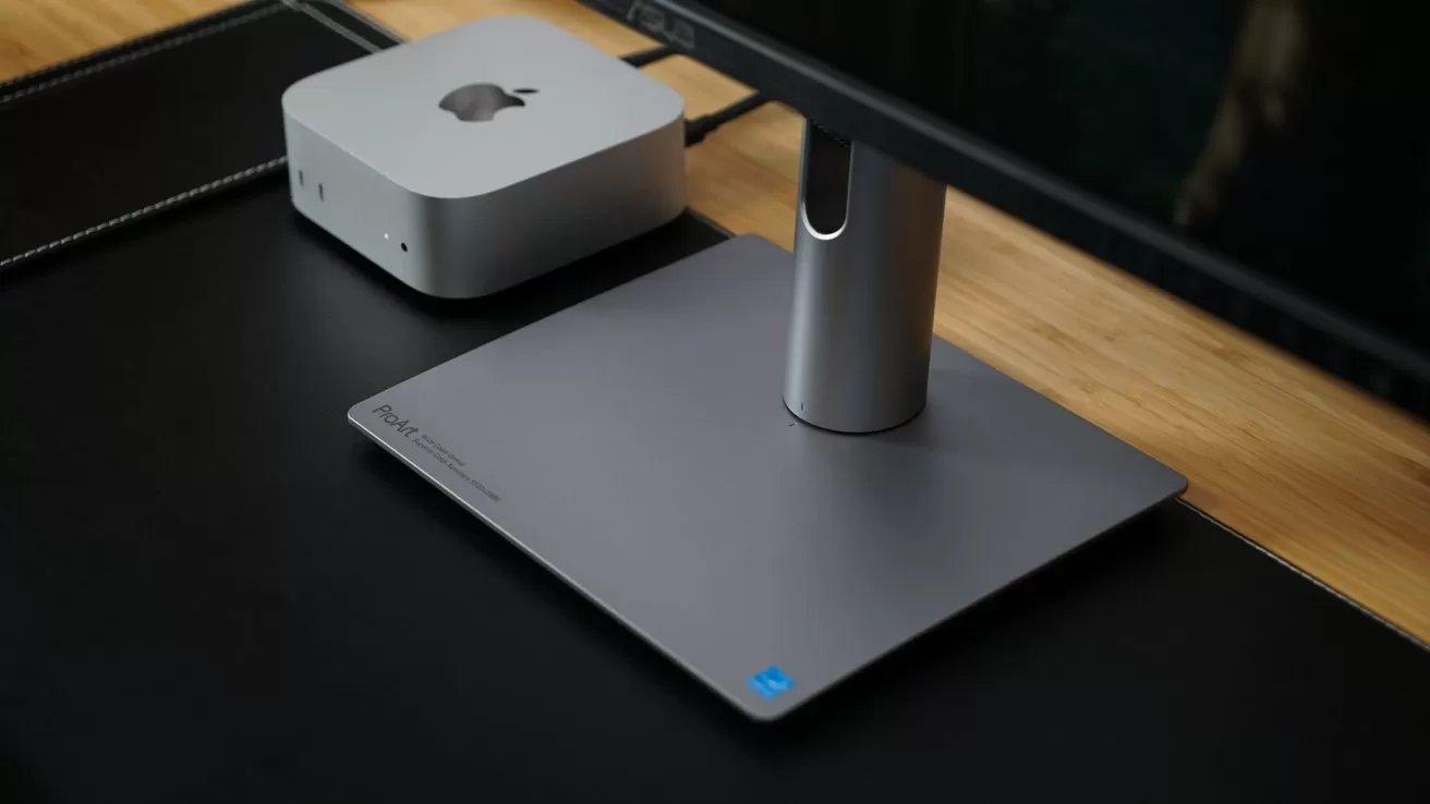 Small silver desktop computer next to a sleek modern monitor stand on a black desk pad, set on a wooden desk in a minimalist workspace