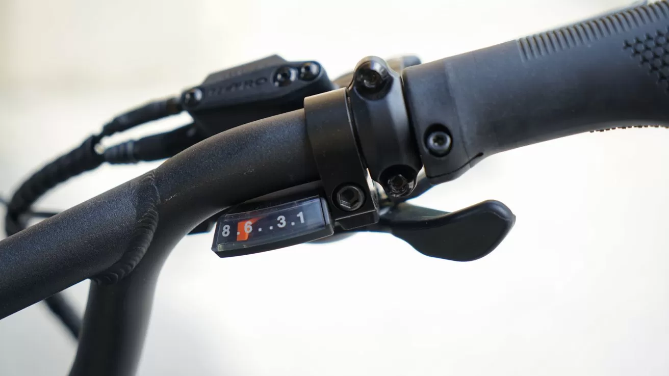 Close-up of a black bicycle handlebar showing textured rubber grip, brake lever, gear shifter, and a small gear indicator window displaying numbers 8, 6, 3, and 1 against a light background