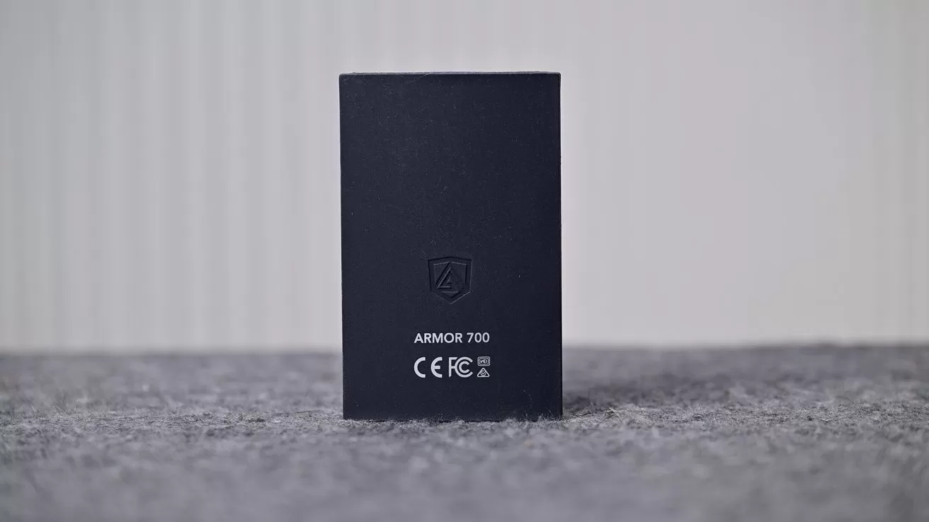 Black rectangular electronic device standing upright on a gray textured surface, showing a shield logo, the text ARMOR 700, and certification symbols against a softly blurred light background