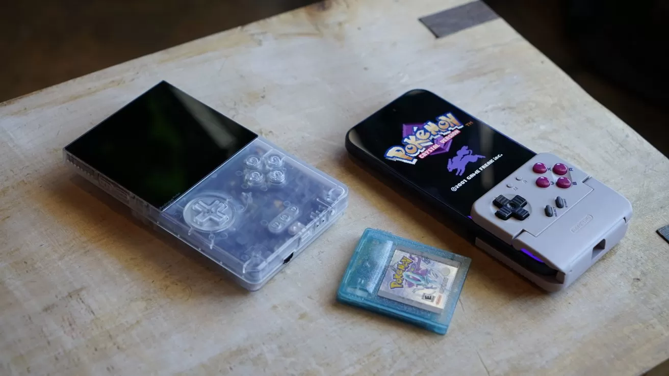 An iPhone, an Analogue Pocket, and a Pokemon game cartridge resting on a wooden surface, including a transparent console and a smartphone-like device with attached retro-style controls